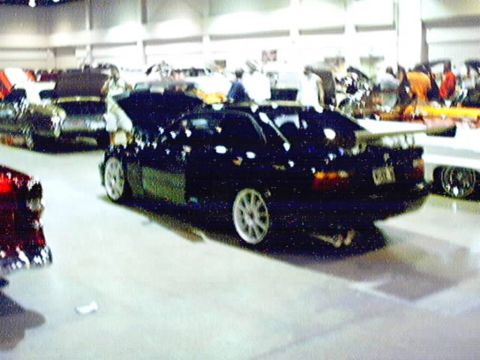 1998 Ford Taurus - A black sedan with a prominent rear spoiler is displayed at a car show, with other vehicles visible in the background.