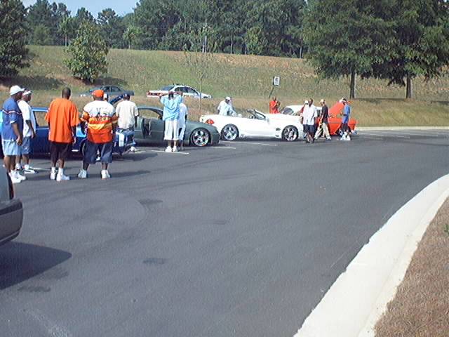 Group of people by a white convertible and a blue sedan at an outdoor gathering