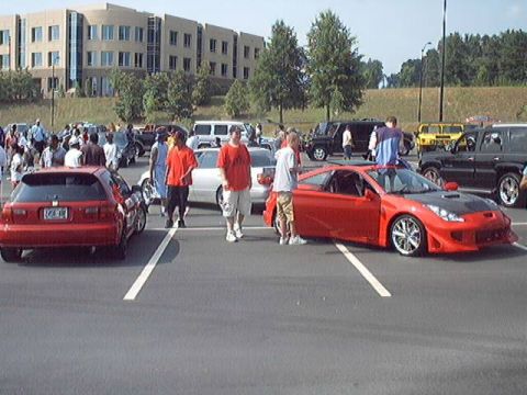 1998 Ford Taurus - A gathering of people and vehicles in a parking lot, a red sports car and a red hatchback.