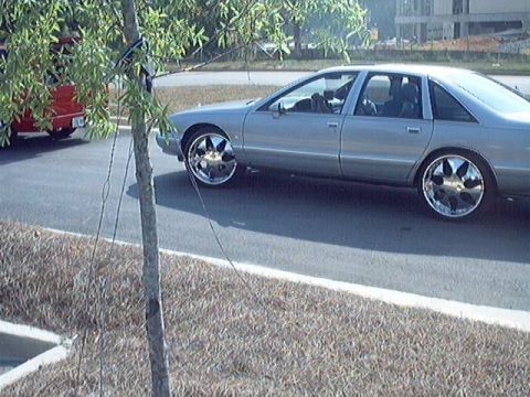 1998 Ford Taurus - A silver sedan with custom chrome rims is parked on the side of the road, with another red vehicle visible in the background.