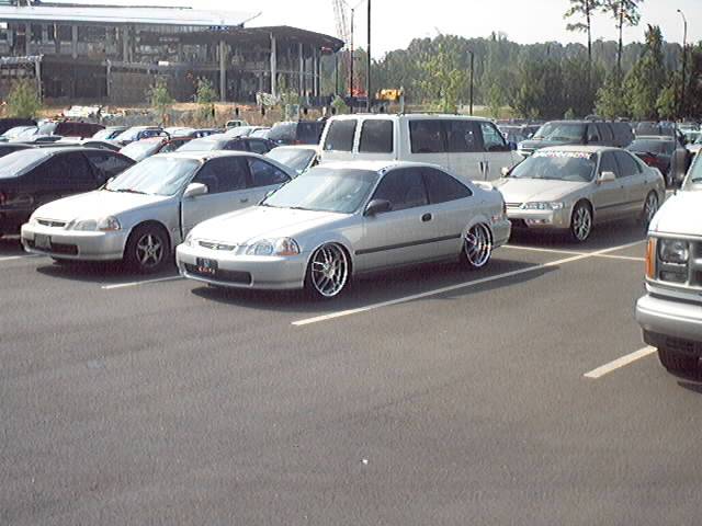Silver two-door coupe with chrome rims parked in a lot, other cars and a van visible