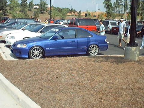 1998 Ford Taurus - A blue coupe is parked in a lot with several other vehicles and pedestrians visible in the background.