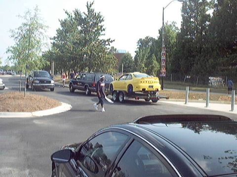1998 Ford Taurus - A yellow coupe is transported on a trailer, pulled by a dark SUV, with a person walking in the street.