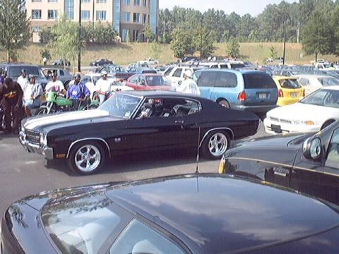 1998 Ford Taurus - A black muscle car with racing stripes is parked in a lot full of vehicles and onlookers.