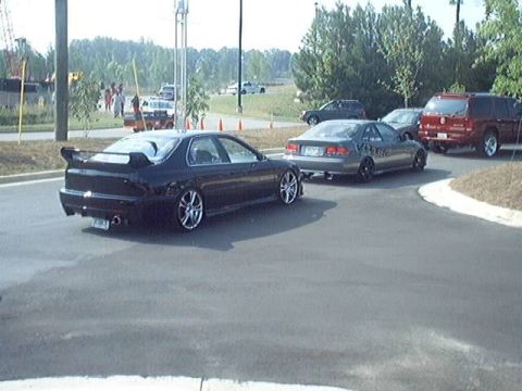 1998 Ford Taurus - Several cars are parked on asphalt, including a black sedan with a spoiler and a red SUV.