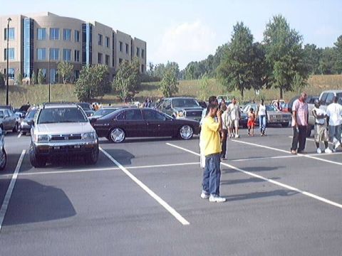 1998 Ford Taurus - A dark sedan with custom chrome wheels is parked in a lot with other vehicles and people.