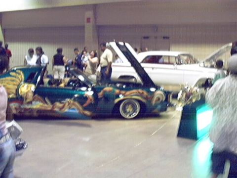 1998 Ford Taurus - A customized teal lowrider is displayed with its hood up, alongside other vehicles at a car show.