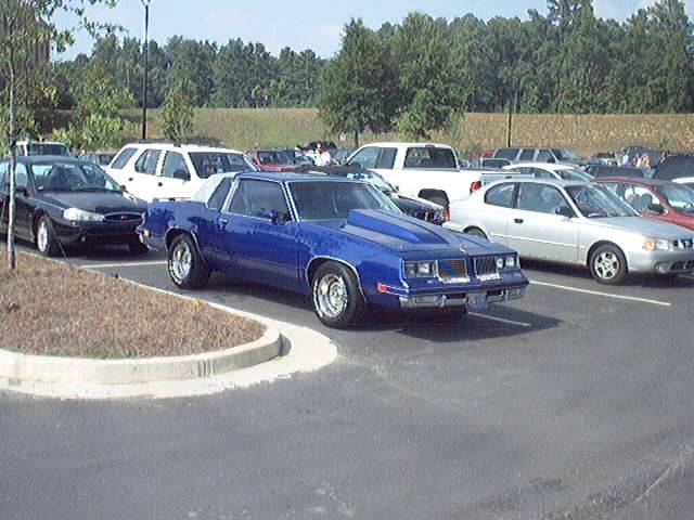 Blue 1980s G-body coupe with hood scoop and chrome wheels in a parking lot