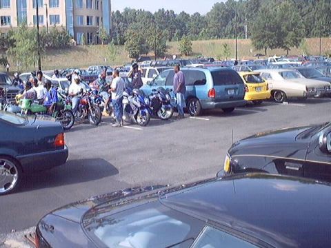 1998 Ford Taurus - A group of people stand around several motorcycles in a large parking lot, with cars and a building visible.