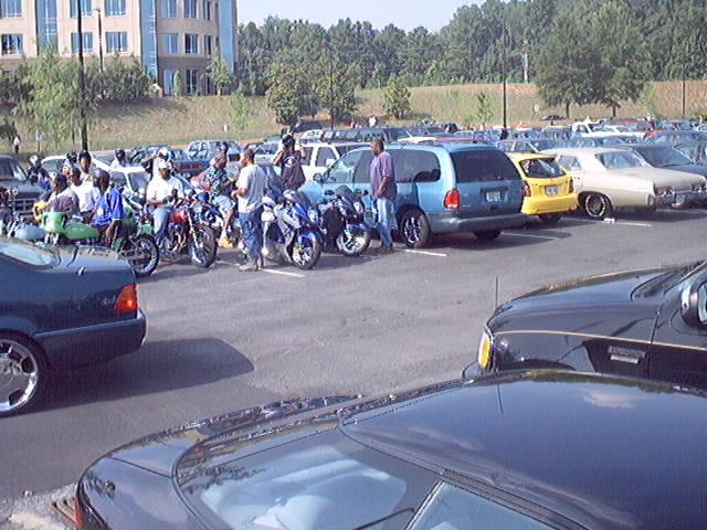 group of people with motorcycles in a parking lot with cars and a building in background