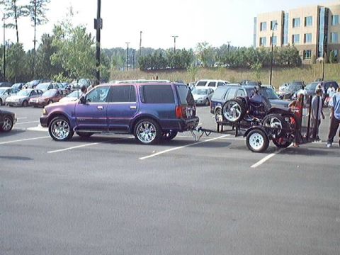 1998 Ford Taurus - A purple SUV is seen towing a trailer carrying a custom vehicle with oversized wheels at a car show.