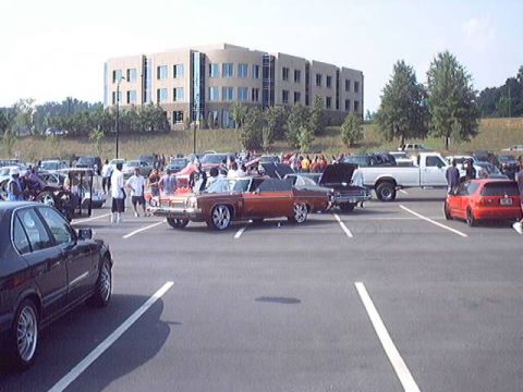 1998 Ford Taurus - A crowd gathers around a classic orange car with custom chrome rims at an outdoor car show.
