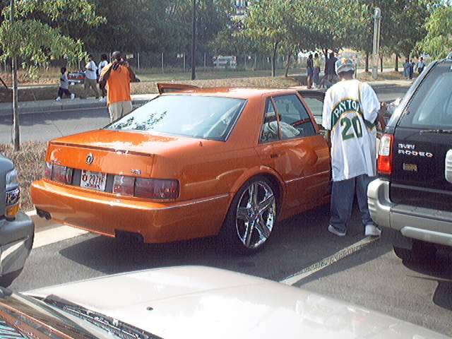 Orange sedan with chrome rims parked next to a black SUV. A person in a white shirt with 'PAYTON 20' is standing between them