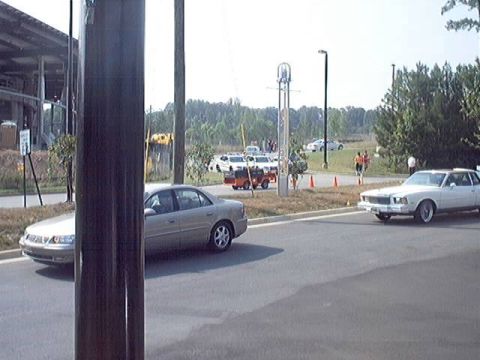 1998 Ford Taurus - Several vehicles are visible on a paved road with a wooded area and buildings in the background.