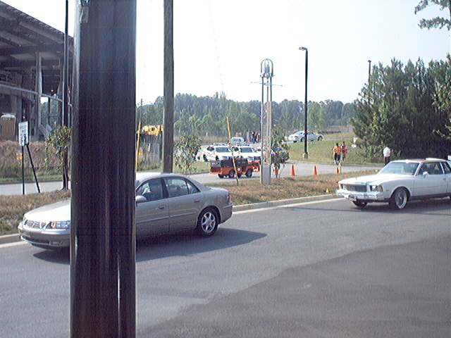 Cars on a road, one is a silver sedan, another is a white coupe. Background has trees and buildings