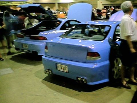 1998 Ford Taurus - Two blue sedans are parked side-by-side at an indoor car show.