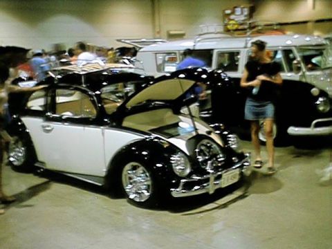 1998 Ford Taurus - A classic Volkswagen Beetle is displayed at a car show, with a woman standing nearby and other vehicles in the background.