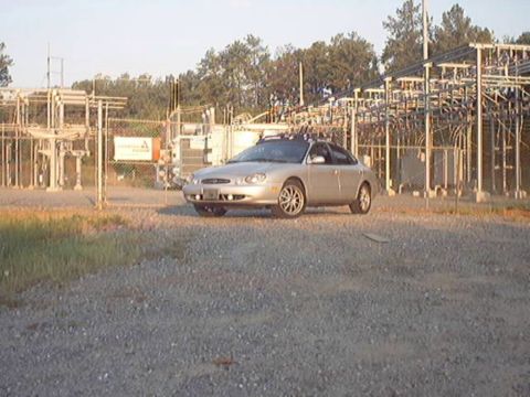 1998 Ford Taurus - Gold/champagne Ford Taurus sedan parked on a gravel lot in front of a fenced electrical substation, shot from a low three-quarter front ang…