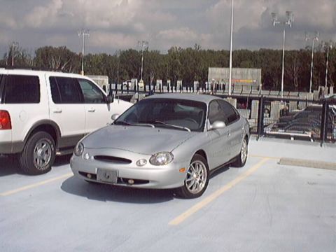 1998 Ford Taurus - Silver four-door sedan with rounded oval-themed styling parked on the top deck of a parking garage, seen from the front three-quarter with…