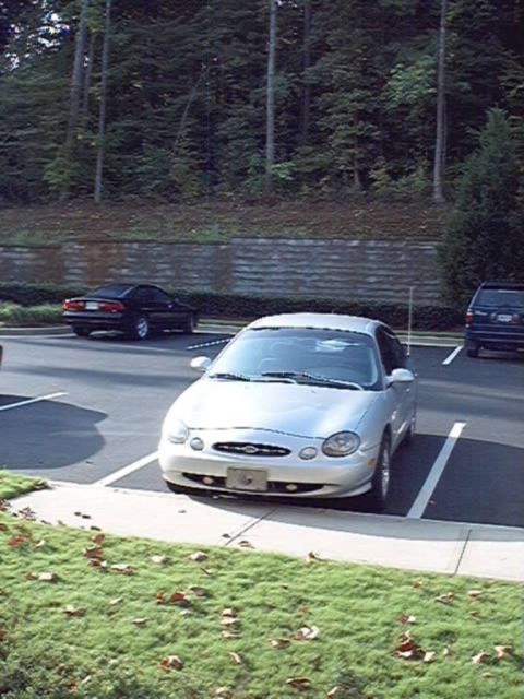 1998 Ford Taurus - Silver oval-styled Ford Taurus sedan parked nose-in in a lot, shot from an elevated front three-quarter angle with a retaining wall and woo…