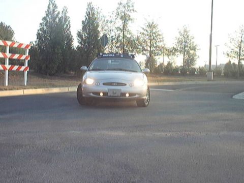 1998 Ford Taurus - Silver Ford Taurus sedan viewed head-on with headlights and fog lights on and a roof-mounted light bar, sitting on a paved lot beside a str…
