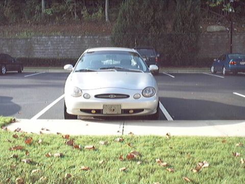 1998 Ford Taurus - Head-on view of a silver four-door sedan nosed into a parking space beside a leaf-strewn grass median, showing the oval-badged grille, roun…