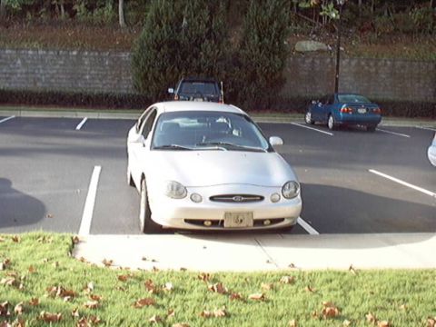 1998 Ford Taurus - Front view of a silver Ford Taurus sedan parked nose-out in a paved lot, showing the oval grille, hood, and windshield; a blue coupe and a…
