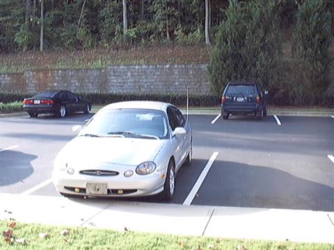 1998 Ford Taurus - Silver oval-era Ford Taurus sedan parked nose-out in a parking lot backed by a stone retaining wall and trees, with a black coupe and a dar…