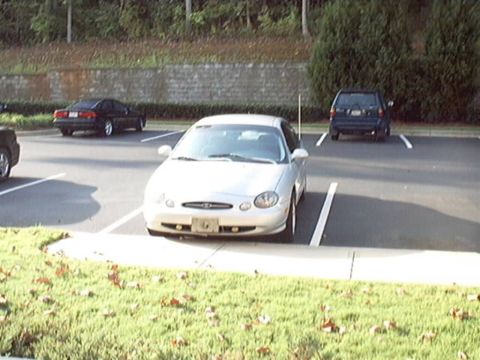 1998 Ford Taurus - Silver Ford Taurus sedan facing the camera, parked nose-out in a lot beside a landscaped bank and stone retaining wall, showing its oval gr…