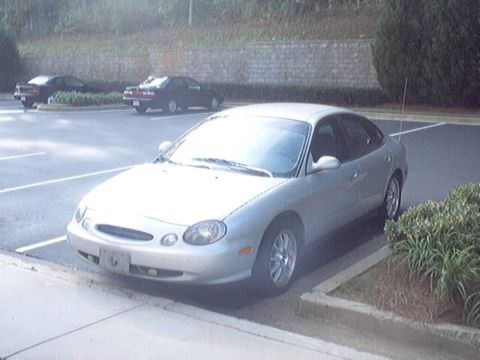 1998 Ford Taurus - Front three-quarter view of a silver oval-era Ford Taurus sedan parked in a lot beside a stone retaining wall and shrubs. It has alloy whee…