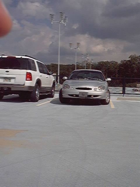 1998 Ford Taurus - A silver Ford Taurus sedan parked nose-out on a rooftop parking deck beside a white Ford Explorer SUV under a cloudy sky.
