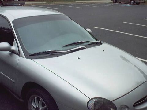 1998 Ford Taurus - Close 3/4 front view of a silver Ford Taurus sedan parked in an empty lot, showing the rounded hood, raked windshield with wipers, driver's…