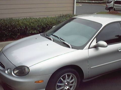 1998 Ford Taurus - High-angle front three-quarter view of a silver Ford Taurus sedan with oval headlights and aftermarket multi-spoke alloy wheels, parked in…