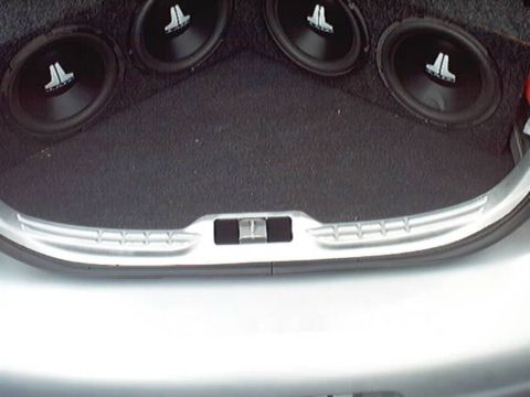 1998 Ford Taurus - Open trunk of a silver sedan with a custom black-carpeted stereo install: four JL Audio subwoofers mounted in an angled enclosure wrapping…