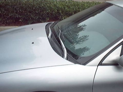 1998 Ford Taurus - Close-up of the cowl and windshield area of a silver sedan, showing a pair of polished chrome wiper arms parked on the glass and two small…