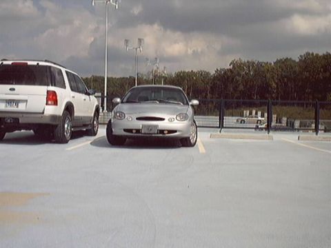 1998 Ford Taurus - Silver Ford Taurus sedan shot head-on from low angle on a white-painted parking deck under a cloudy sky. The car sits low on dark wheels be…