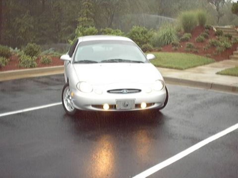 1998 Ford Taurus - A silver Ford Taurus sedan photographed head-on with its headlights and fog lights illuminated, parked on a wet asphalt lot in the rain wit…
