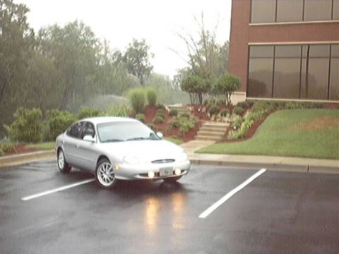1998 Ford Taurus - A silver Ford Taurus sedan parked at an angle across the striped stalls of a wet office parking lot, headlights and fog lights on in the ra…