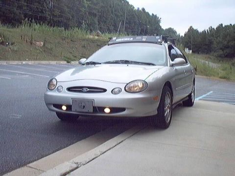 1998 Ford Taurus - Front three-quarter view of a silver oval-era Ford Taurus sedan parked along a curb with fog lights lit, fitted with a roof rack with wind…
