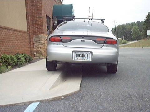 1998 Ford Taurus - Rear view of a silver Ford Taurus sedan with a roof rack, parked on a driveway beside a brick building. The vanity license plate reads 'NOS…