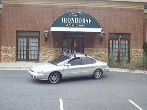 1998 Ford Taurus - Silver four-door Ford Taurus sedan parked at an angle in an empty lot in front of a brick building with a green awning reading "Ironhorse o…