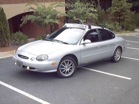 1998 Ford Taurus - Front three-quarter view of a silver Ford Taurus sedan fitted with a roof rack with a wind fairing and bike-carrier mounts, plus multi-spok…