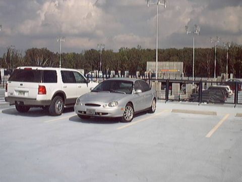1998 Ford Taurus - Silver Ford Taurus sedan parked beside a white Ford Explorer SUV on an open rooftop parking deck under a cloudy sky. The Taurus is seen fro…