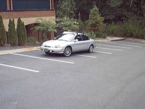 1998 Ford Taurus - Silver Ford Taurus four-door sedan parked alone in an empty striped asphalt lot, shot from a distant 3/4 front angle. It wears multi-spoke…