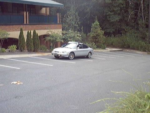 1998 Ford Taurus - Silver four-door sedan parked alone in an empty striped asphalt lot, shot from a distance at a front three-quarter angle. A brick-and-tan t…