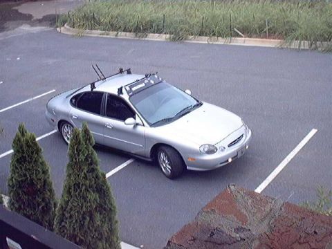 1998 Ford Taurus - Elevated three-quarter front view of a silver late-1990s Ford Taurus sedan parked across spaces in an empty lot, fitted with a roof rack ca…