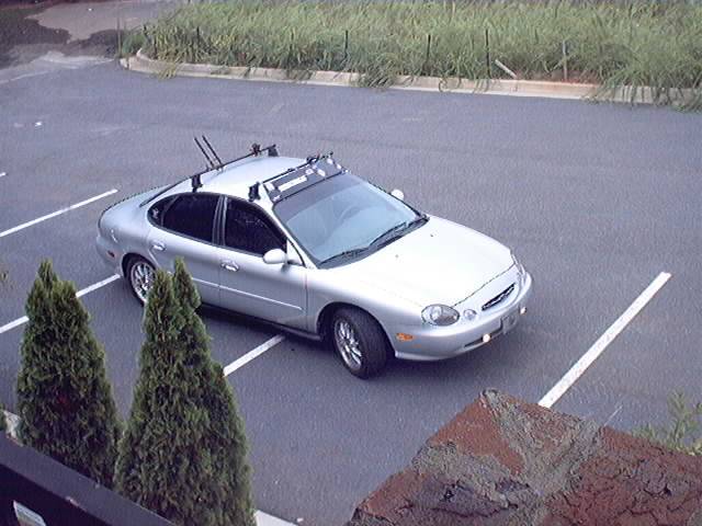 Silver Ford Taurus sedan with roof rack and bike carriers, viewed from above in an empty parking lot
