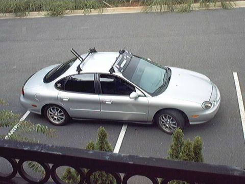 1998 Ford Taurus - High-angle view of a silver Ford Taurus sedan parked across painted parking-space lines, fitted with aftermarket roof-rack crossbars and ca…