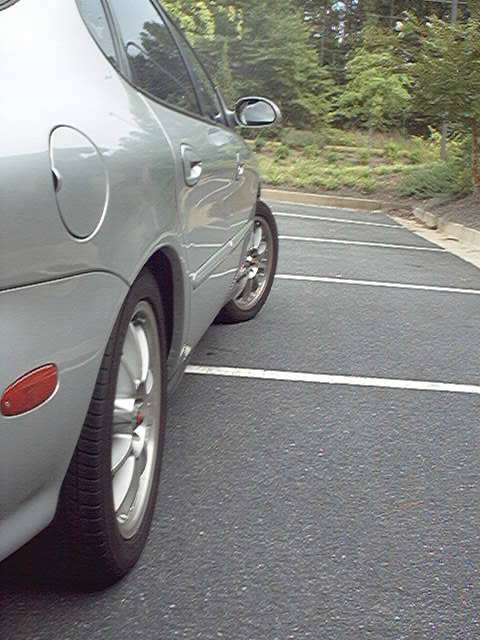 1998 Ford Taurus - Low-angle view down the passenger side of a silver sedan parked in an empty lot, shot from behind the rear wheel and looking forward along…