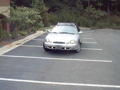 1998 Ford Taurus - Silver Ford Taurus sedan photographed head-on in an empty striped asphalt parking lot at dusk, backed by a wooded treeline and brick buildi…
